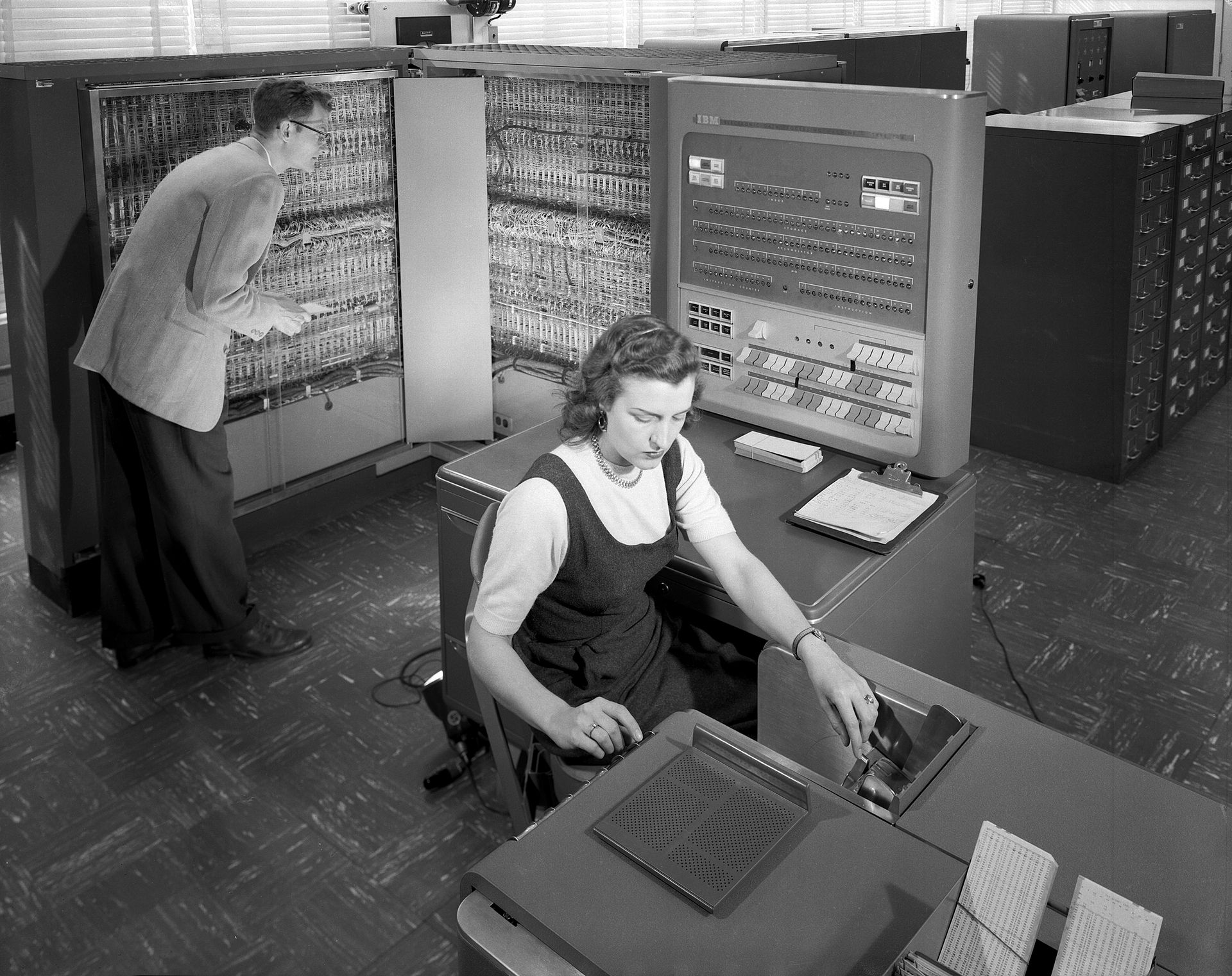 An image of an IBM mainframe in the background of a room with two persons working on it. One person is wearing a suit and looking at the mainframe panel. The other person is wearing a dress and looks to be typing at a terminal or other interface.